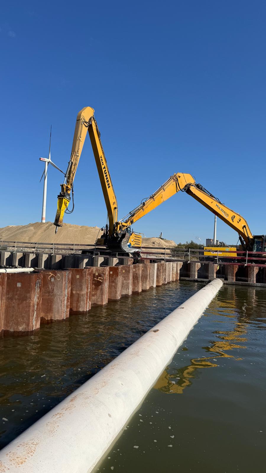 Twee rupskranen met lange giek langs een damwand, met een zandberg en een windturbine op de achtergrond