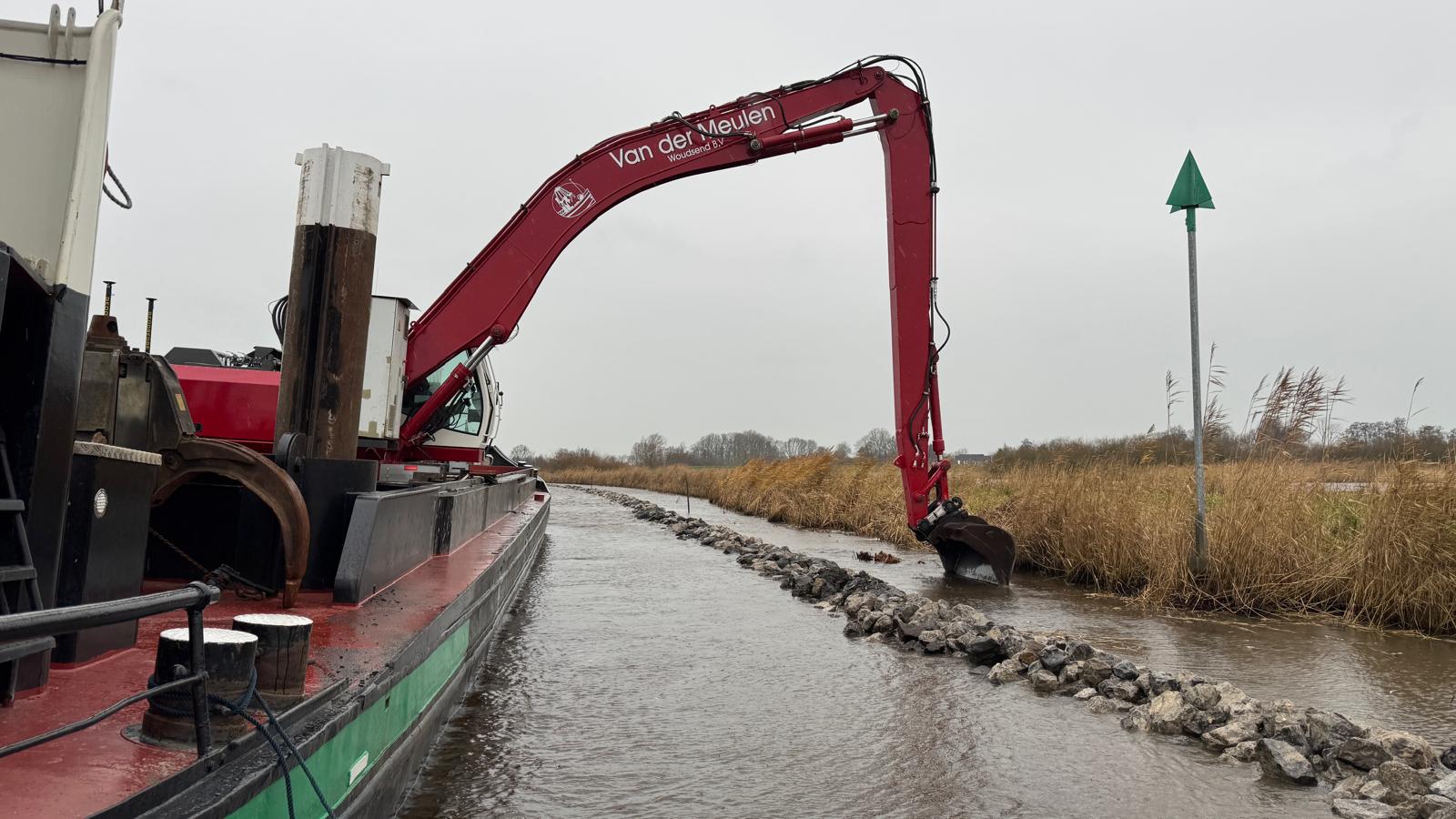 Langarmgraafmachine baggert een vaarweg langs een stenen oever, met een baggerschip aan de linkerkant