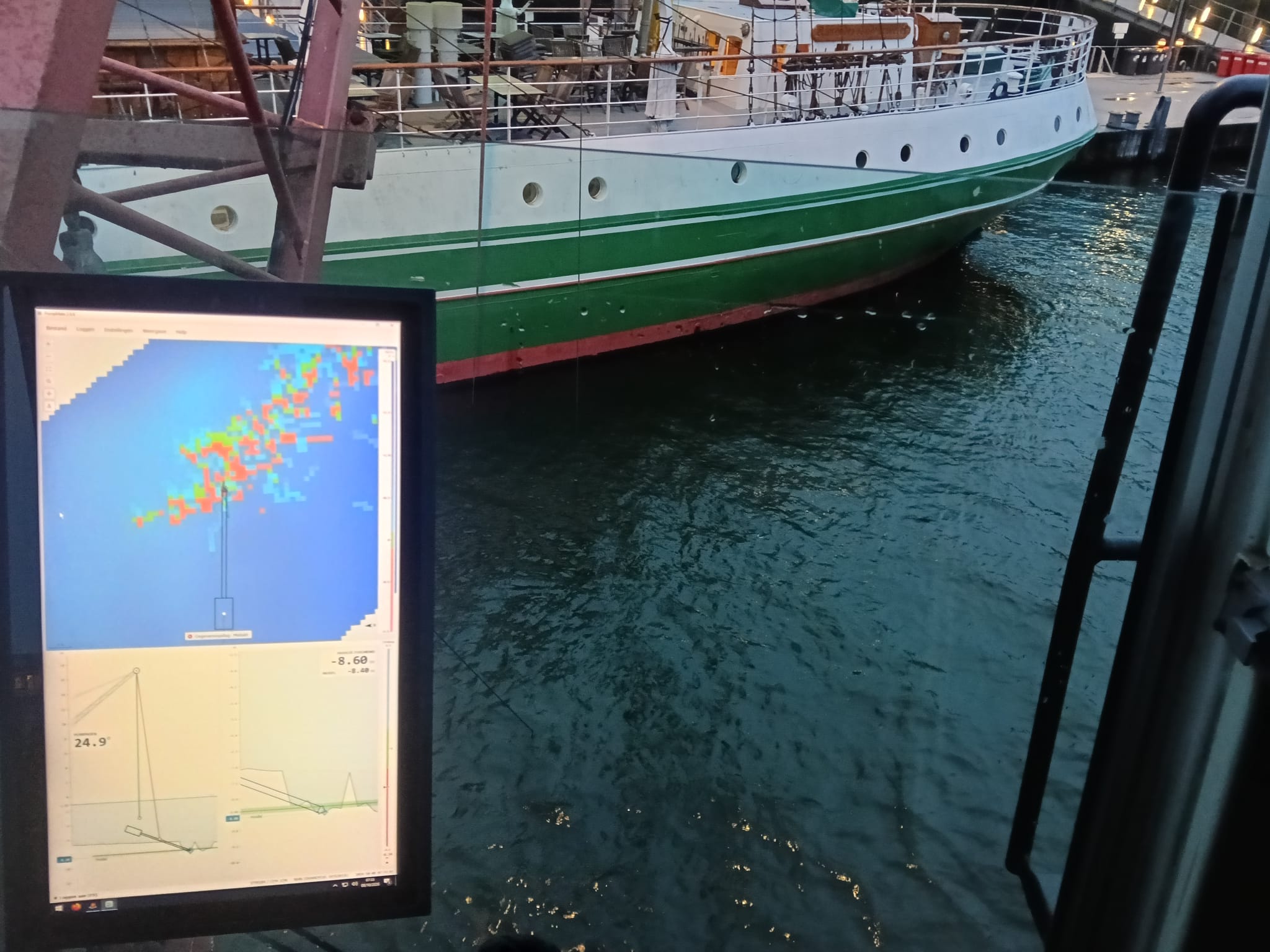 Scherm in de cabine met positie- en ontgravingsdata, met een schip aan de kade op de achtergrond